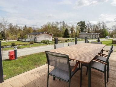 An outdoor patio with a wooden table and chairs overlooking a gravel road and wooden cabins at 4 Meadow Retreat in Dobwalls