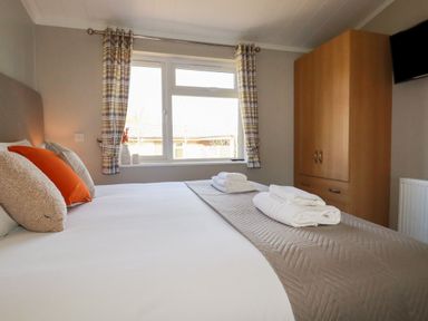 A bedroom with a made bed with towels on it a window with curtains a wooden wardrobe and a wall mounted tv at 23 Meadow Retreat in Dobwalls