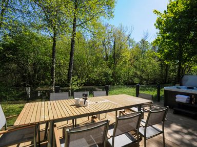 An outdoor wooden dining table with chairs on a deck surrounded by trees and a hot tub nearby at 23 Meadow Retreat in Dobwalls