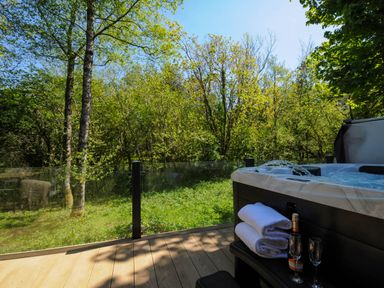 An outdoor deck with a hot tub towels bottle and glasses overlooking trees and grass at 23 Meadow Retreat in Dobwalls