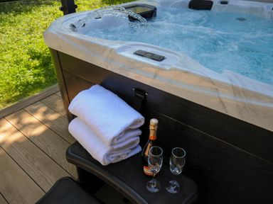 A hot tub with water flowing in next to folded towels and a bottle with two glasses on a wooden deck at 23 Meadow Retreat in Dobwalls