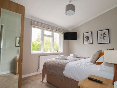 A bedroom with a bed with folded towels on it a bedside table with a lamp and remote control a wall-mounted television a wardrobe with a mirror and windows with checked curtains at 6 Faraway Fields Dobwalls