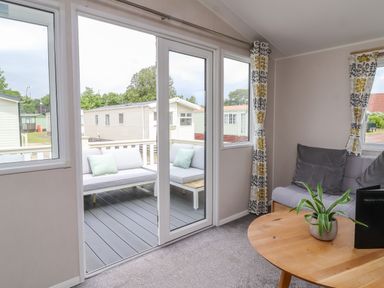 A living room with a sofa a wooden table and a plant next to glass doors leading to an outdoor seating area at Windermere Lodge in Morecambe