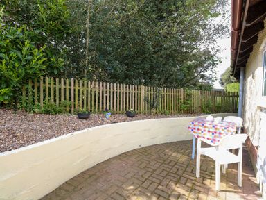 An outdoor patio area with a small table covered by a checkered tablecloth and two white chairs next to a house with a wooden fence and plants in the background at Paws-A-While in Hoveton & Wroxham