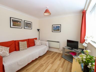 A living room with a red sofa, a small TV on a stand, framed pictures on the walls, and flowers on a table at Paws-A-While in Hoveton & Wroxham