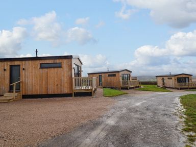 Three wooden cabins with small porches on gravel and grassy land under a partly cloudy sky at Cae Pren in New Mills near Llanfair Caereinion