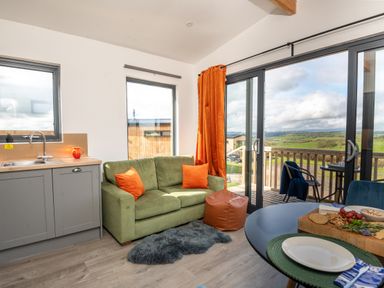 A living room with a green sofa and orange cushions next to a kitchen sink and large glass doors opening to a balcony with countryside view at Cae Pren in New Mills near Llanfair Caereinion