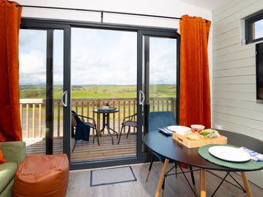 A dining area with a black table set for two with food on the table and a sliding glass door leading to a balcony with two chairs and a small round table with a view of green fields at Cae Pren in New Mills near Llanfair Caereinion