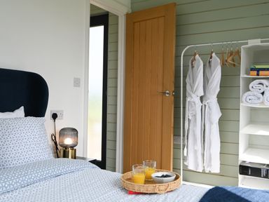 A bedroom with a bed a bedside lamp a door robes on a rack and shelves with towels and books at Cae Pren in New Mills near Llanfair Caereinion