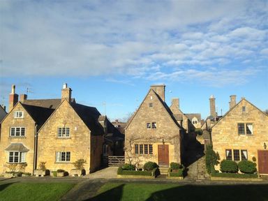 Three stone houses with chimneys and small gardens in front under a partly cloudy sky in Broadway