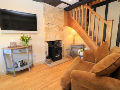 A living room with a brown sofa a stone fireplace a wood burning stove a wall mounted television a wooden staircase a console table with flowers and books at Broadway Cottage in Broadway
