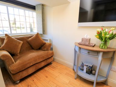 A brown couch with two cushions next to a window and a small blue table with books flowers and tissue box in a room at Broadway Cottage in Broadway