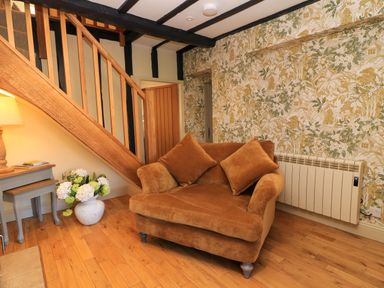 A sitting area with a brown sofa and cushions a wooden staircase side tables a lamp and floral wallpaper at Broadway Cottage in Broadway