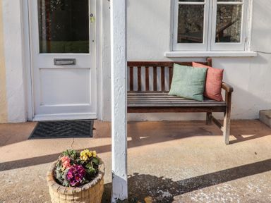 A porch with a wooden bench with cushions next to a white door and a flower pot on the ground at 4 Lymbrook Cottages in Lyme Regis