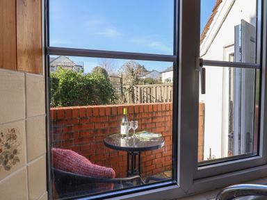 A table with a bottle of wine, two glasses, and a book on a small patio with a chair outside a window at 4 Lymbrook Cottages in Lyme Regis