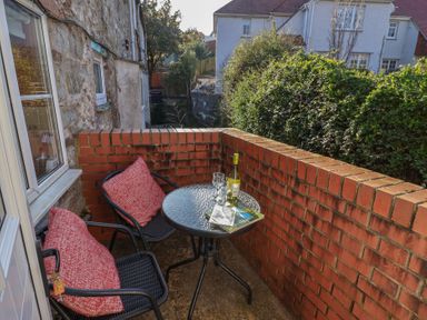 A small balcony with two chairs with red cushions a round glass table with a bottle of wine two glasses and brochures at 4 Lymbrook Cottages in Lyme Regis