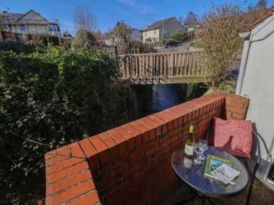 A small outdoor balcony with a round glass table holding a bottle of wine two wine glasses and books near a brick wall and a wooden bridge over a stream at 4 Lymbrook Cottages Lyme Regis