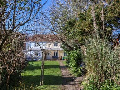 A grassy garden with a dirt path leading to a two-story white house surrounded by trees and bushes at 4 Lymbrook Cottages in Lyme Regis