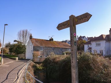 A wooden signpost indicating directions to Lyme Regis and Uplyme by a path near houses and a small water channel at 4 Lymbrook Cottages in Lyme Regis