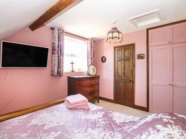 A bedroom with pink walls a wall-mounted television a wooden dresser a wooden door and a bed with floral bedding at The Old Chapel in Foxley near Bawdeswell