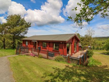 A red wooden cabin with a fenced patio and outdoor seating surrounded by grass and trees at Moonlake Lodge in Woolsery near Woolfardisworthy