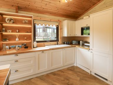 A kitchen with white cabinets wooden countertops a sink under a window and shelves with decorations at Moonlake Lodge in Woolsery near Woolfardisworthy
