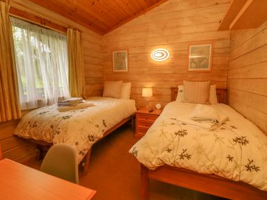 A bedroom with two single beds a window with curtains a small bedside table with a lamp and framed pictures on wooden walls at Moonlake Lodge in Woolsery near Woolfardisworthy