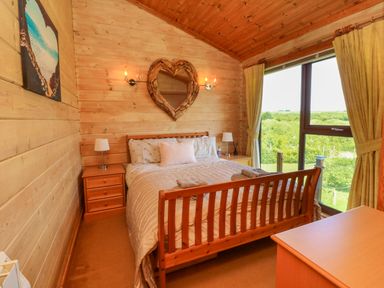 A bedroom with a wooden bed and side tables next to a large window showing green trees at Moonlake Lodge in Woolsery near Woolfardisworthy