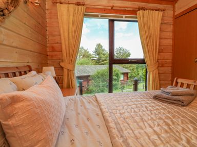 A bedroom with a bed facing a window with curtains showing trees and a wooden building outside at Moonlake Lodge in Woolsery near Woolfardisworthy