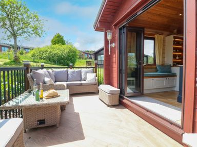 An outdoor patio with a wicker sofa and table next to an open doorway leading to a wooden interior at Moonlake Lodge in Woolsery near Woolfardisworthy