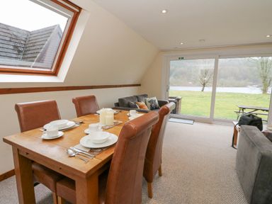 A dining area with a wooden table set with dishes and four brown chairs next to a living room with sofas and a large glass door overlooking a grassy yard at Lodge 2 - Inglewood Lodge in Spean Bridge