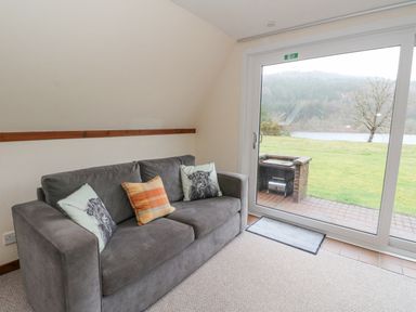A living room with a gray sofa and cushions near a large glass door overlooking a lawn and lake at Lodge 2 - Inglewood Lodge in Spean Bridge