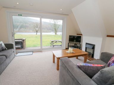 A living room with two gray sofas a wooden coffee table a television and a fireplace with large sliding glass doors overlooking grass and trees at Lodge 2 - Inglewood Lodge in Spean Bridge