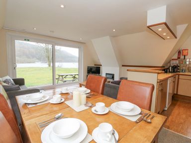 A dining table set with plates and cups in an open living room and kitchen area overlooking a lawn and lake through large sliding glass doors at Lodge 2 - Inglewood Lodge in Spean Bridge