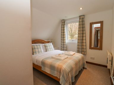 A bedroom with a double bed with plaid bedding and two pillows a window with plaid curtains a wooden framed mirror and a bedside lamp at Lodge 2 - Inglewood Lodge in Spean Bridge
