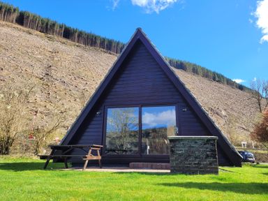 An a frame cabin with glass doors next to a wooden picnic table on grass with a cleared hillside in the background at Lodge 2 - Inglewood Lodge in Spean Bridge