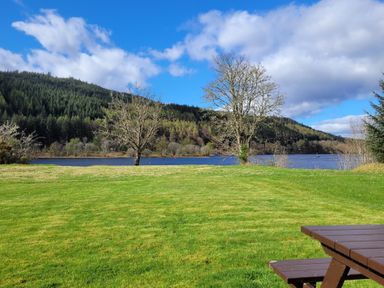 A grassy area with trees near a lake and a wooden picnic table at Lodge 2 - Inglewood Lodge in Spean Bridge