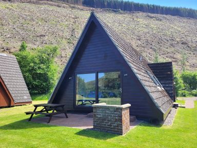 An A-frame cabin with glass sliding doors and a wooden picnic table on a grassy area at Lodge 2 - Inglewood Lodge in Spean Bridge