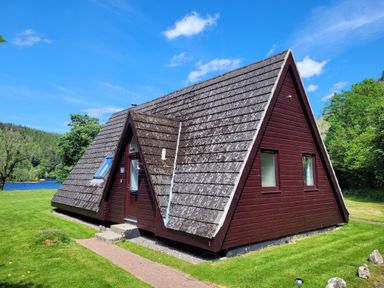 An A-frame wooden cabin with a shingled roof near a grassy area and trees by a lake at Lodge 2 - Inglewood Lodge in Spean Bridge