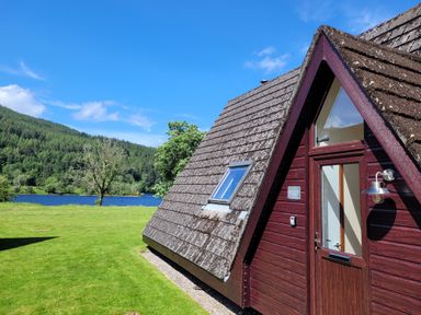 A wooden triangular lodge with a door and window near a lake and forest at Lodge 2 - Inglewood Lodge in Spean Bridge