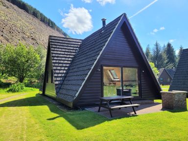 A triangular cabin with a picnic table and brick barbecue on grass near a hillside at Lodge 2 - Inglewood Lodge in Spean Bridge