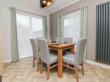 A dining room with a wooden table and six grey chairs near windows with green patterned curtains at 18 Manleigh Park in Combe Martin