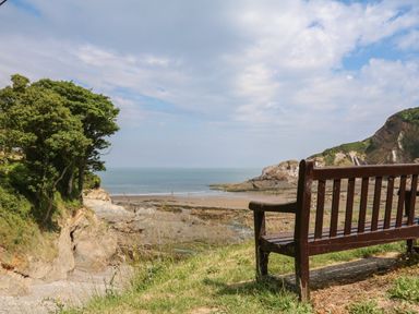 A wooden bench overlooking a rocky beach sea and trees at 18 Manleigh Park in Combe Martin