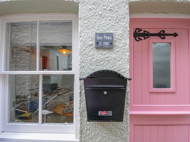 A window and a pink door with a black mailbox and a sign saying Sea Pinks c 1780 on a white textured wall at Sea Pinks in Looe