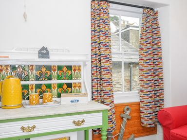 A room corner with a green and white cabinet with yellow kettle and cups next to a window with fish patterned curtains at Sea Pinks in Looe