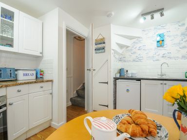 A kitchen with white cabinets a table with croissants and a teapot and a staircase in the corner at Sea Pinks in Looe