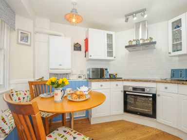 A kitchen with a round wooden table, chairs with patterned cushions, white cabinets, a built-in oven, microwave, and a vase with yellow flowers at Sea Pinks in Looe