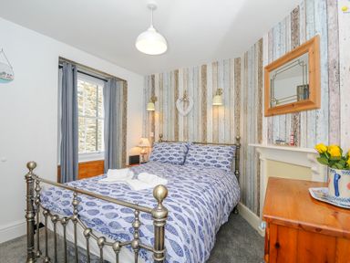 A bedroom with a metal bed frame blue patterned bedding a wooden chest of drawers and striped wallpaper at Sea Pinks in Looe