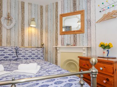 A bedroom with a metal bed with fish patterned bedding a wooden dresser with yellow flowers a mirror and striped wallpaper at Sea Pinks in Looe