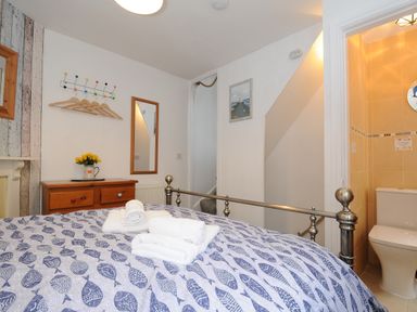 A bedroom with a metal bed frame a blue fish patterned bedspread a wooden dresser with a vase of yellow flowers a mirror and an adjoining bathroom at Sea Pinks in Looe